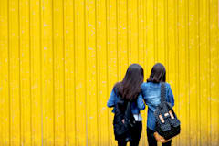 两位黑发女孩背包离摄像头远望黄墙